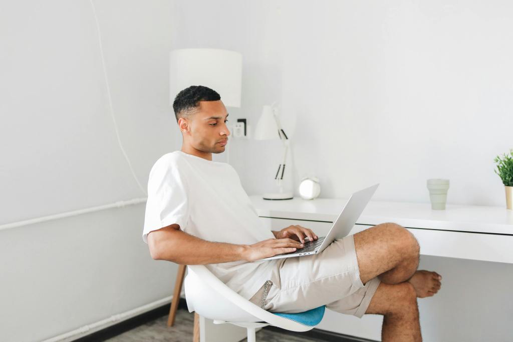 A man sitting in a modern chair at a desk, working on a laptop in a minimalist room.